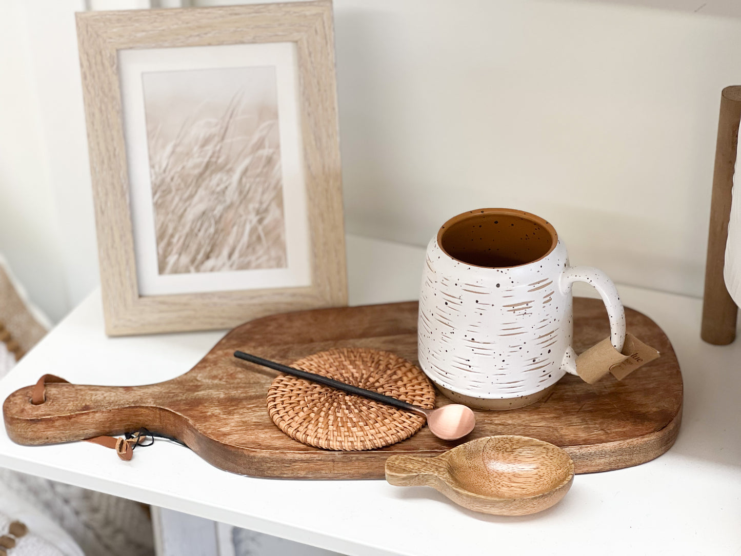 An eggshell white handled coffee mug sitting on a wooden cutting board. Added accessories include a coaster, copper stir spoon, and a mango wood scoop spoon. The stoneware mug is white in color with a light scratch pattern to it that allows a small dimension and contrasting tan color to peak through. 