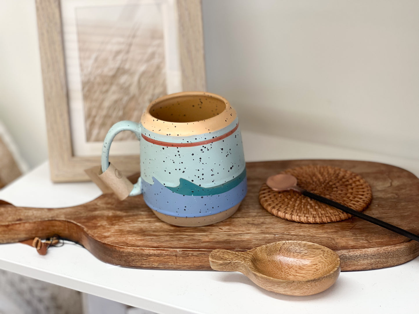 A multi-colored (blue, aqua, light blue, white, rust, tan) handled coffee mug sitting on a wooden cutting board. Added accessories include a coaster, copper stir spoon, and a mango wood scoop spoon. The stoneware mug is multi-colored with a wave pattern on it.