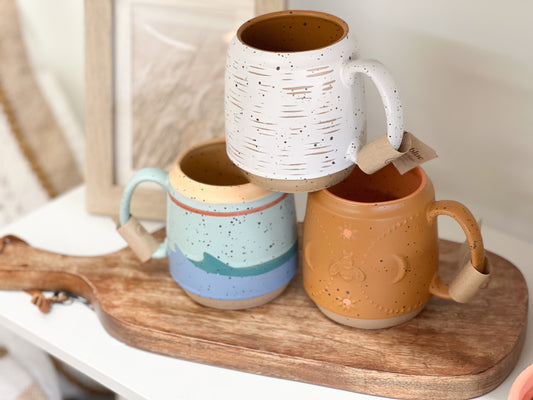 3 stoneware coffee mugs stacked into a pyramid. They are boho in style. The top mug is eggshell white with a scratch pattern, the bottom right is orange or terracotta color with a celestial pattern, and the bottom left is a multi color (blue, orange, and white) mug with a wave pattern 