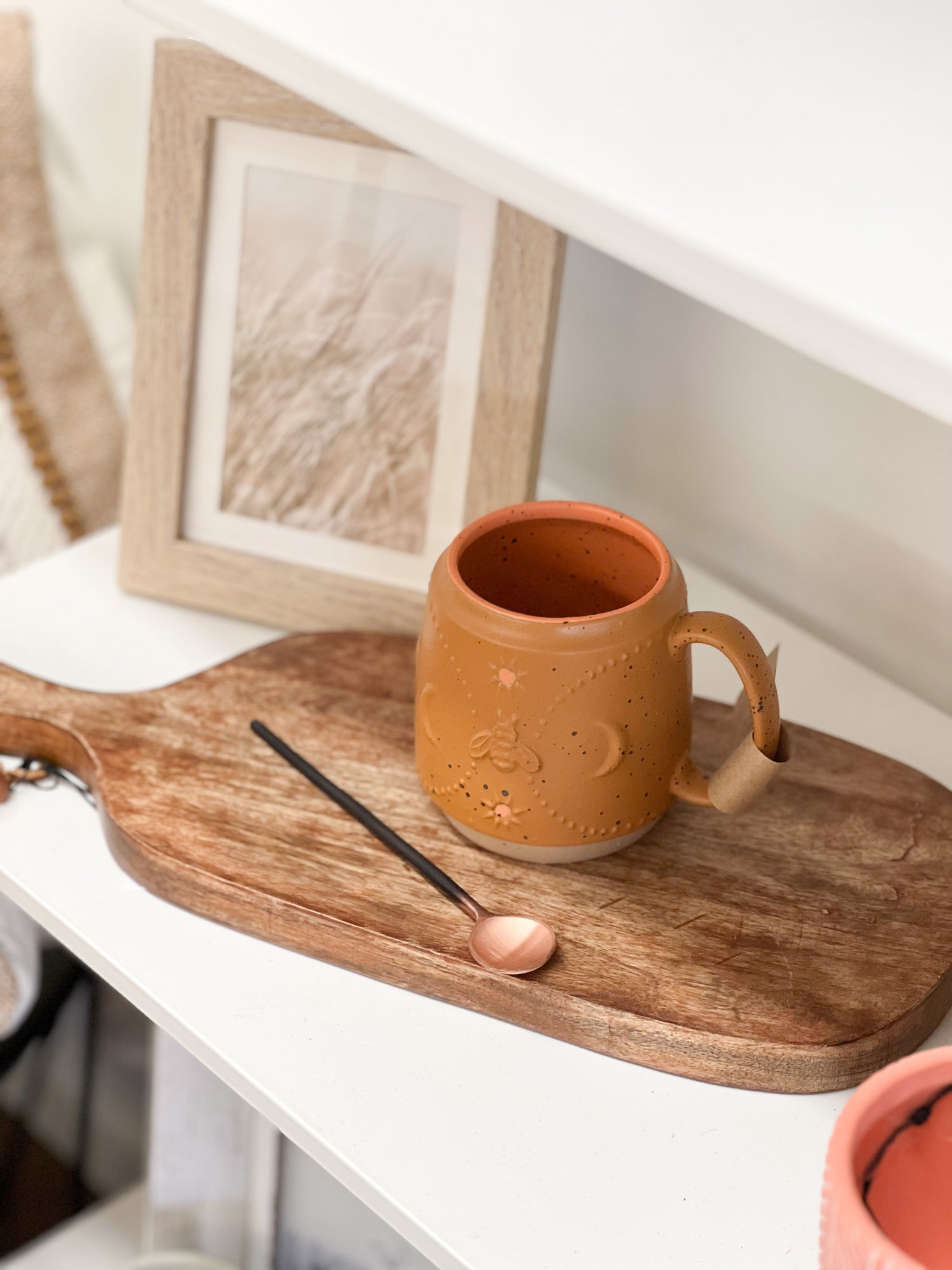 A terracotta handled coffee mug sitting on a wooden cutting board. Added accessories include copper stir spoon, and a light wood picture frame in the background. The stoneware mug is terracotta in color with a celestial pattern detail wrapped around the surface.